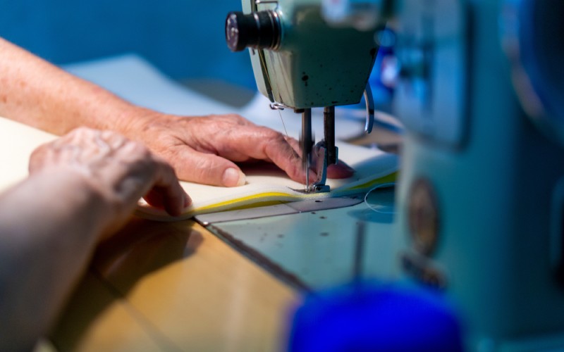 Two hands holding a large piece of white fabric on top of yellow fabric under the needle of an industrial sewing machine