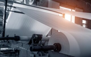 Textile manufacturing machine producing a large roll of white fabric stretched through various tubes inside an industrial environment.